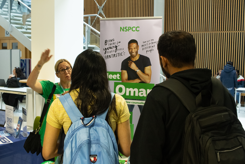 Image is of two students stood talking to a charity representative at the Volunteer Fair